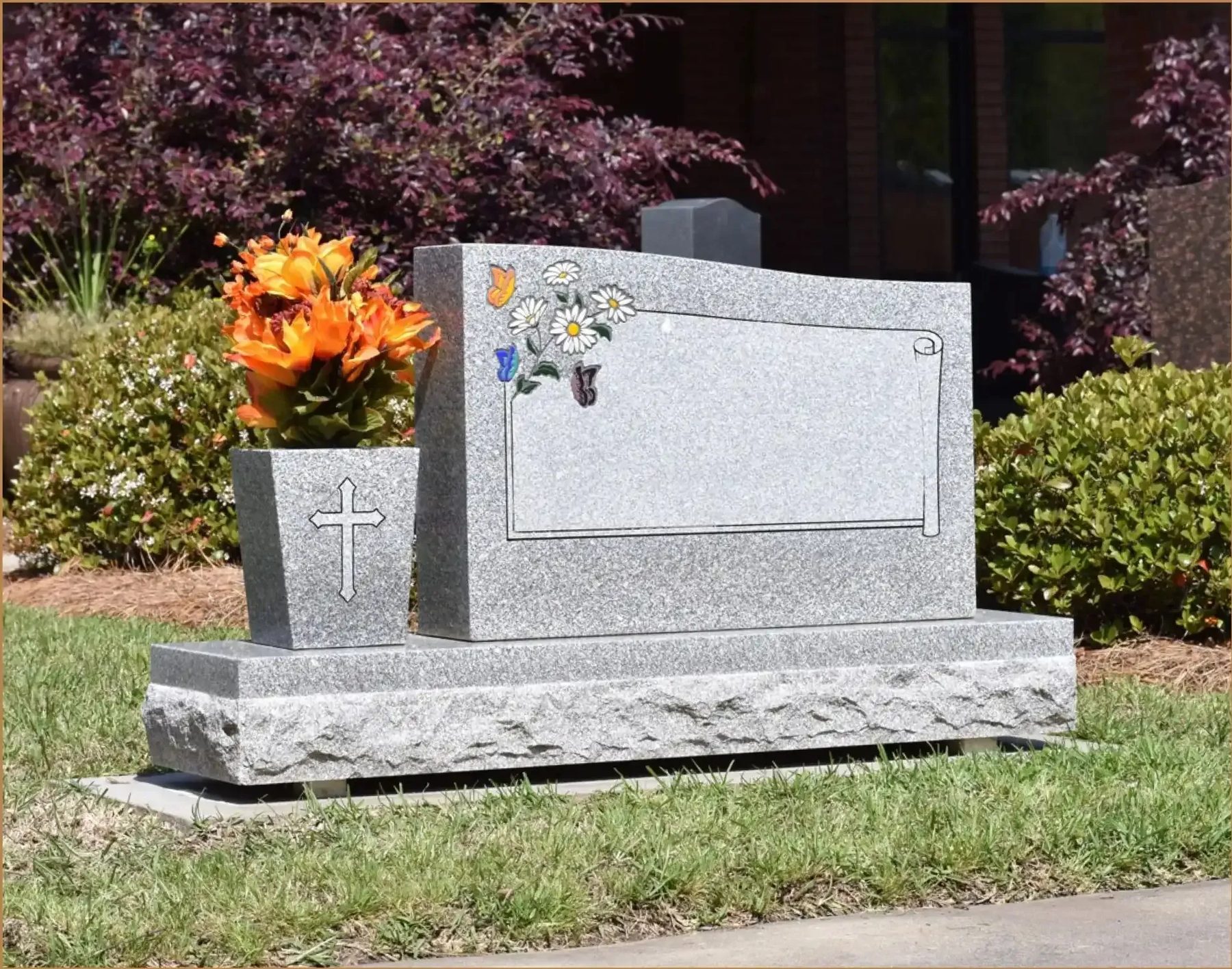A grey granite companion monument with a carved scroll panel, daisies and butterflies, standing on a rock-faced base in a cemetery