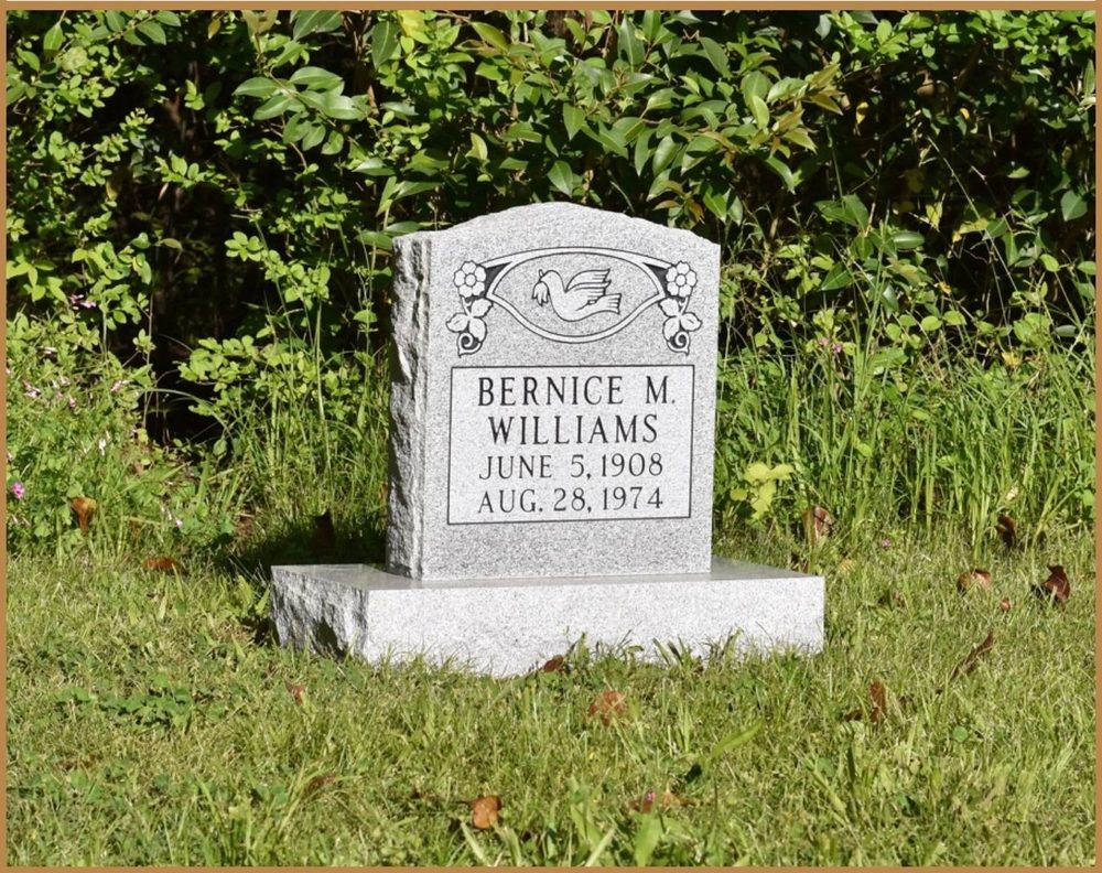 A small upright granite monument with a shaped top standing in grass