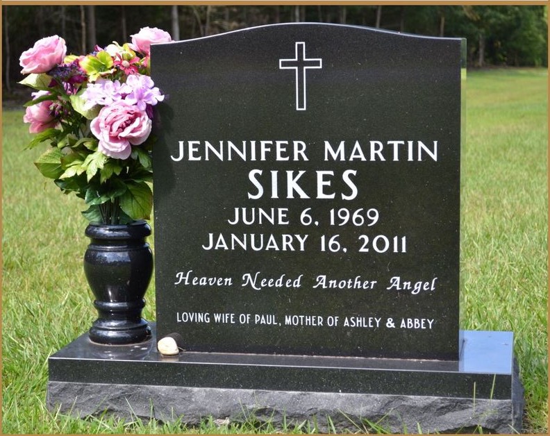 A dark granite upright monument with a carved cross and flowers at its base
