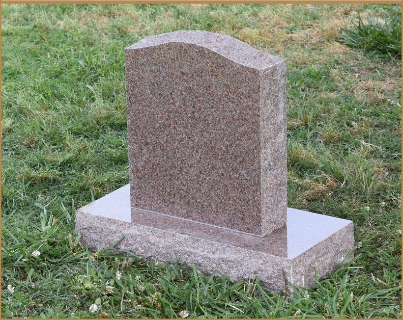A rose granite upright monument with a shaped top, standing in grass